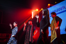 A group of performers on stage wearing dark outfits and scarves, holding raised hands in a synchronized dance pose