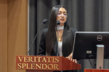 A person standing at a lectern with the inscription ‘Veritatis Splendor’, next to a computer monitor. The speaker is addressing an audience in a lecture hall with curtains in the background.”