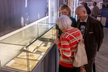 Visiting a museum exhibition. Three people stand by a glass display case and observe historical documents and dried plant specimens labeled with exhibition tags.