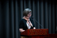 Kobieta stoi przy mównicy z napisem „Veritatis Splendor” i czyta z przygotowanych materiałów. Przed kobietą znajdują się dwa mikrofony.
