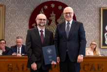 Dwóch mężczyzn stoi obok siebie w sali senatu WUM. Mężczyzna po lewej trzyma zamkniętą teczkę z dokumentem. Obaj mężczyźni uśmiechają się do zdjęcia. W tle siedzą uczestnicy posiedzenia oraz widoczny jest herb uczelni.