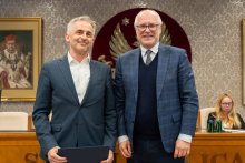 Dwóch mężczyzn stoi obok siebie w sali senatu WUM. Mężczyzna po lewej trzyma zamkniętą teczkę z dokumentem. Obaj mężczyźni uśmiechają się do zdjęcia. W tle siedzą uczestnicy posiedzenia oraz widoczny jest herb uczelni.