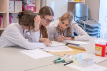 Trzy osoby w fartuchach laboratoryjnych siedzą przy stole i pracują z dokumentami. Jedna osoba wskazuje palcem fragment tekstu, dwie pozostałe analizują treść i notują. Na stole leżą kartki, długopis, telefon oraz sprzęt laboratoryjny.