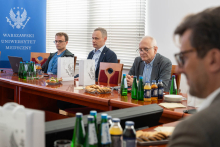 Kilku mężczyzn siedzi przy konferencyjnym stole podczas spotkania na Warszawskim Uniwersytecie Medycznym. Mężczyźni mają przed sobą laptopy, butelki wody, szklanki oraz talerze z przekąskami, a w tle znajduje się roll‑up z logo Warszawskiego Uniwersytetu Medycznego.