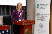 Kobieta osoba stoi przy mównicy i przemawia do mikrofonu. Obok znajduje się ekran projekcyjny oraz baner z nazwą konferencji logopedii klinicznej.