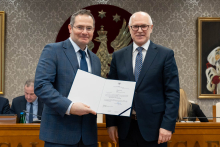 Dwóch mężczyzn stoi obok siebie. Ten po lewej prezentuje otwarty dokument. Obaj mężczyźni uśmiechają się. a nimi widoczny jest herb herb uczelni - złoty orzeł na czerwonym tle oraz sala obrad. 