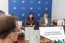 Kobieta i mężczyzna siedzą przy stole konferencyjnym na tle ściany z powtarzającym się logo Warszawskiego Uniwersytetu Medycznego. Na stole stoją butelki wody, napojów oraz torby uczelniane, a przed uczestnikami znajduje się ekran z napisem „Konwent Pracodawców Wydziału Lekarskiego WUM – II posiedzenie”.