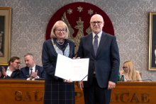 Kobieta i mężczyzna stoją obok siebie w sali senatu WUM. Kobieta trzyma otwartą teczkę z dokumentem. Obie osoby uśmiechają się do zdjęcia. W tle siedzą uczestnicy posiedzenia oraz widoczny jest herb uczelni.