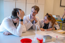 Trzy osoby w fartuchach laboratoryjnych siedzą przy stole i omawiają zadanie. Jedna osoba opiera głowę na dłoni, druga patrzy na rozmówcę, trzecia pochyla się nad dokumentami. Na stole leżą kartki i pojemniki laboratoryjne.