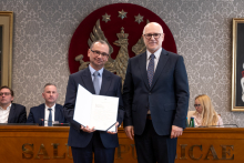 Dwóch mężczyzn stoi obok siebie w sali senatu WUM. Mężczyzna po lewej trzyma otwartą teczkę z dokumentem. Obaj mężczyźni uśmiechają się do zdjęcia. W tle siedzą uczestnicy posiedzenia oraz widoczny jest herb uczelni.