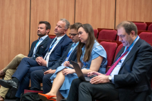 Uczestnicy konferencji siedzą w rzędzie czerwonych foteli w sali wykładowej. Mężczyźni i kobieta mają identyfikatory na smyczach, jedna osoba trzyma laptop na kolanach, a pozostali opierają dłonie na kolanach lub trzymają notatki.