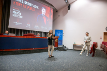 Jedna osoba stoi na scenie i przemawia do mikrofonu trzymanego w ręce. Obok, przy schodach, inna osoba stoi i obserwuje wydarzenie. W tle widoczny jest duży ekran z informacjami o wykładzie „Profesjonalny język, który nie poniża” oraz data „25 maja 2026” i godzina „17:00”.