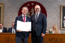 Dwóch mężczyzn stoi obok siebie w sali senatu WUM. Mężczyzna po lewej trzyma otwartą teczkę z dokumentem. Obaj mężczyźni uśmiechają się do zdjęcia. W tle siedzą uczestnicy posiedzenia oraz widoczny jest herb uczelni.