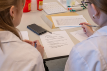Dwie osoby w fartuchach laboratoryjnych siedzą obok siebie przy stole i czytają dokument z zadaniami. Jedna osoba trzyma długopis i robi notatki. Na stole leżą kartki, telefon oraz drobny sprzęt laboratoryjny.