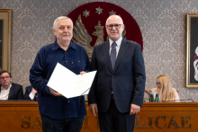 Dwóch mężczyzn stoi obok siebie w sali senatu WUM. Mężczyzna po lewej trzyma otwartą teczkę z dokumentem. Obaj mężczyźni uśmiechają się do zdjęcia. W tle siedzą uczestnicy posiedzenia oraz widoczny jest herb uczelni.