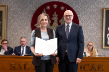 Kobieta i mężczyzna stoją obok siebie w sali senatu WUM. Kobieta trzyma otwartą teczkę z dokumentem. Obie osoby uśmiechają się do zdjęcia. W tle siedzą uczestnicy posiedzenia oraz widoczny jest herb uczelni.