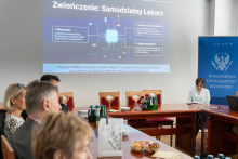 Uczestnicy spotkania siedzą przy stole konferencyjnym i patrzą na rzutnik, na którym wyświetlana jest prezentacja pod tytułem „Zwieńczenie: Samodzielny Lekarz”. Na stole stoją butelki wody, torby uczelniane oraz talerze z poczęstunkiem, a obok znajduje się banner Warszawskiego Uniwersytetu Medycznego.