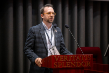 Osoba w okularach stoi przy mównicy z napisem „VERITATIS SPLENDOR” i przemawia do mikrofonów. Na szyi ma identyfikator konferencyjny, a ręce opiera o pulpit podczas wystąpienia.
