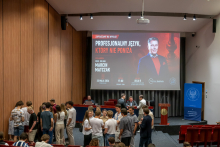 Grupa kilkunastu osób stoi i rozmawia w sali wykładowej przed sceną. W tle widoczny jest duży ekran z plakatem wydarzenia zawierającym napis „Profesjonalny język, który nie poniża” oraz datę i miejsce wykładu. Na scenie znajduje się mównica z napisem „VERITATIS SPLENDOR”.