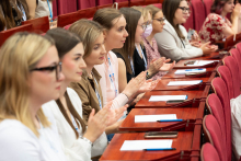 Osiem osób siedzi w rzędzie i klaszcze. Przed nimi znajdują się pulpity z kartkami i długopisami.