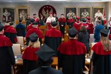 Kilkadziesiąt osób ubranych w tradycyjne tego akademickie stoi w dużej sali. Na ścianach wiszą portrety, jest też godło WUM - złoty orzeł na czerwonym tle.