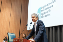 Mężczyzna stoi przy mównicy i przemawia. Za nim siedzą dwie osoby przy stole prezydialnym, a na ekranie wyświetla się tytuł konferencji.