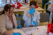 Kilka osób w fartuchach laboratoryjnych pracuje przy stołach. Na pierwszym planie jedna osoba używa pipety, druga obserwuje jej działania. Na stołach stoją pojemniki na odpady medyczne i zestawy do pracy laboratoryjnej.