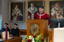Dziekan ubrany w togę akademicką stoi przy mównicy i przemawia. Za nią siedzą dwie osoby. Na ścianie wiszą 3 portrety w ozdobnych ramach.