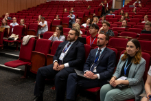 Uczestnicy konferencji siedzą na czerwonych fotelach w auli i słuchają wystąpienia. Na pierwszym planie siedzą mężczyźni i kobieta w formalnych strojach, którzy trzymają notatniki lub mają identyfikatory na smyczach.