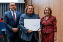 Dwóch mężczyzn i kobieta pozują razem do zdjęcia. Mężczyzna pośrodku trzyma w rękach otwarty dyplom.