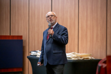 Osoba w garniturze stoi przy stole i przemawia do mikrofonu ręcznego. W tle widoczny jest drewniany panel ścienny oraz stos materiałów lub toreb konferencyjnych leżących na stole.
