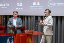 Dwie osoby stoją przy drewnianej mównicy z napisem „VERITATIS SPLENDOR” i prowadzą wystąpienie. Jedna osoba gestykuluje rękami podczas mówienia, druga stoi obok blisko komputera. W tle widoczny jest ekran z informacjami o wydarzeniu, w tym data „25 maja 2026” i godzina „17:00”.