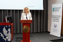 Kobieta stoi przy mównicy i przemawia. Za nią widać baner z nazwą konferencji/