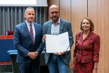 Dwóch mężczyzn i kobieta pozują razem do zdjęcia. Mężczyzna pośrodku trzyma w rękach otwarty dyplom.