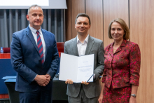 Dwóch mężczyzn i kobieta pozują razem do zdjęcia. Mężczyzna pośrodku trzyma w rękach otwarty dyplom.