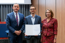 Dwóch mężczyzn i kobieta pozują razem do zdjęcia. Mężczyzna pośrodku trzyma w rękach otwarty dyplom.