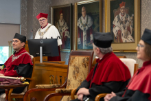 Rektor ubrany w biało-czerwoną togę akademicką stoi przy pulpicie i przemawia do zgromadzonych. Przed nią siedzą cztery osoby w pierwszym rzędzie. W tle wiszą portrety.