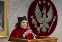 Kobieta ubrana w togę akademicką osoba stoi przy mównicy i mówi do mikrofonu. Za nim widać godło WUM - złotego orła na czerwonym tle.