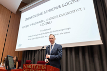 Mężczyzna stoi przy mównicy i przemawia. Na ekranie widnieje tytuł prezentacji dotyczącej neurologopedii.