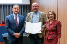 Dwóch mężczyzn i kobieta pozują razem do zdjęcia. Mężczyzna pośrodku trzyma w rękach otwarty dyplom.