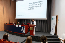 Kobieta stoi przy mównicy i prowadzi prezentację. W tle widoczny jest slajd z ikoną młotka i tekstem dotyczącym regulacji prawnych.