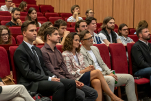Kilkanaście osób siedzi w rzędach czerwonych foteli w sali wykładowej i słucha wystąpienia. W pierwszym rzędzie kilka osób siedzi obok siebie, jedna osoba trzyma w ręce butelkę. W tle widoczne są kolejne rzędy miejsc oraz uczestnicy wydarzenia.