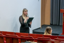 Kobieta stoi między rzędami czerwonych foteli i czyta z zielonej teczki, trzymając mikrofon w dłoni.