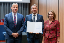 Dwóch mężczyzn i kobieta pozują razem do zdjęcia. Mężczyzna pośrodku trzyma w rękach otwarty dyplom.