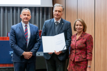 Dwóch mężczyzn i kobieta pozują razem do zdjęcia. Mężczyzna pośrodku trzyma w rękach otwarty dyplom.