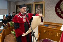 Dziekan stoi przy mikrofonie i odczytuje to, co jest napisane na dużym papirusie. W tle widać inne osoby.