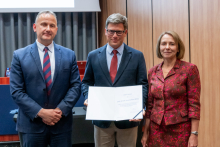 Dwóch mężczyzn i kobieta pozują razem do zdjęcia. Mężczyzna pośrodku trzyma w rękach otwarty dyplom.