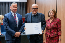 Dwóch mężczyzn i kobieta pozują razem do zdjęcia. Mężczyzna pośrodku trzyma w rękach otwarty dyplom.