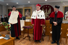 Trzy osoby stoją obok siebie. Jedna osoba trzyma duży dokument. Przed nimi stoi stolik z insygniami.