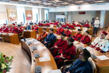 Kilkadziesiąt osób siedzi w ławach i patrzy w stronę mównicy. Wszyscy ubrani są w stroje akademickie.